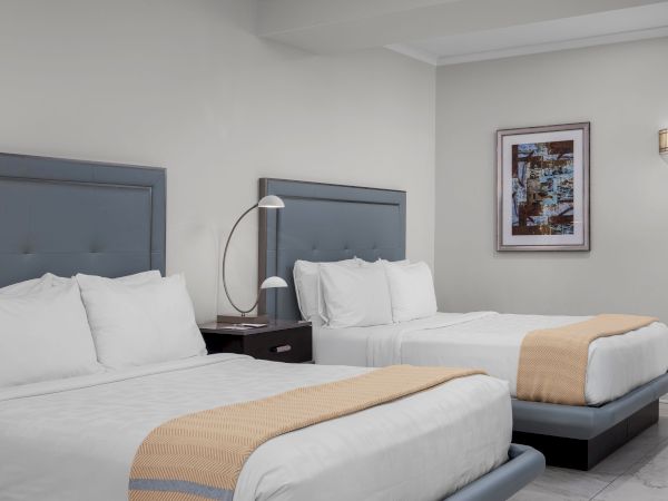 The image shows a hotel room with two beds, white linens, a bedside lamp, art on the wall, and a modern, clean design.