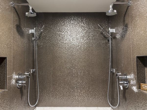 The image shows a modern shower space with two showerheads and handheld sprays, set against a textured wall with built-in shelves.
