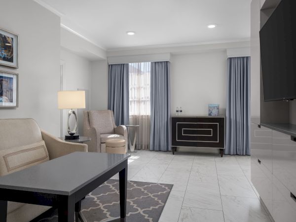 A modern living room with gray curtains, beige sofas, a black coffee table, framed artwork, and a flat-screen TV on the wall.