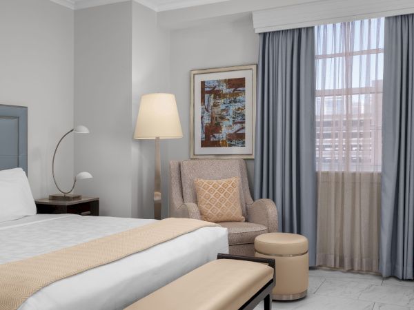 A cozy hotel room with a bed, armchair, lamp, decorative cushion, artwork, and window with blue curtains.