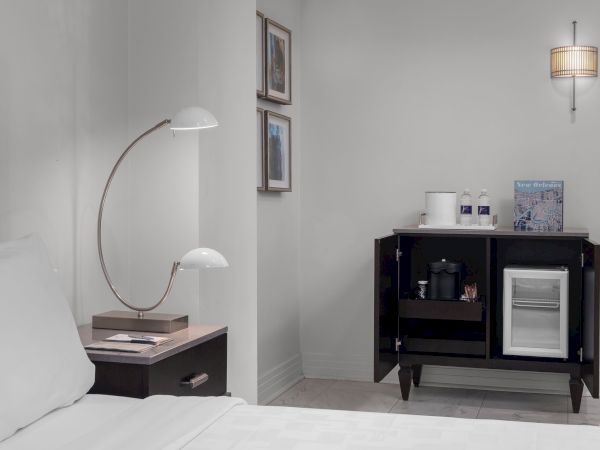 The image shows a hotel room with a bed, a modern lamp, framed art, cabinet, minibar, two water bottles, and a New Orleans map.