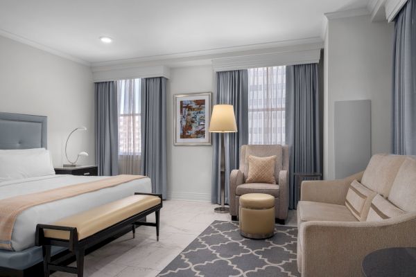 The image shows a modern hotel room with a bed, armchair, sofa, and decor featuring curtains and a floor lamp.