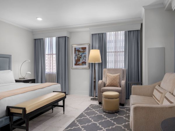 The image shows a modern hotel room with a bed, armchair, sofa, and decor featuring curtains and a floor lamp.