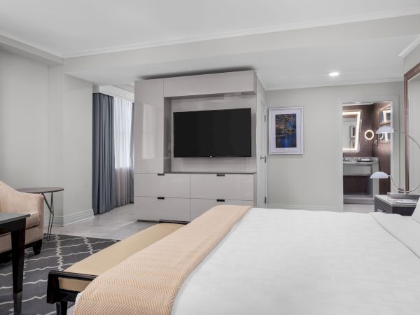 A modern hotel room with a bed, TV, table, armchair, and bathroom with a lighted mirror in view.