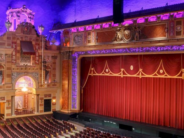 The image shows an ornate theater with a red curtain, intricate architecture, and tiered seating under a purple-lit ceiling.