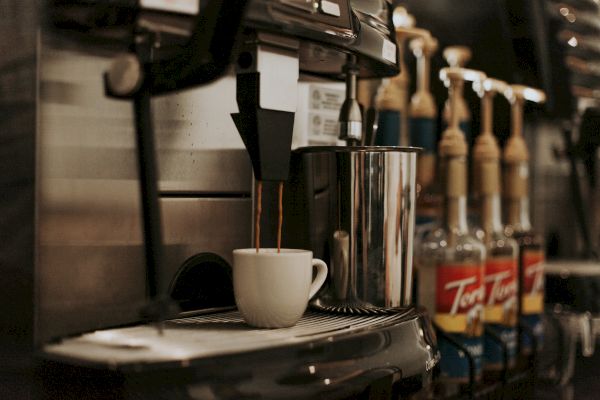 A coffee machine fills a cup, with various syrup bottles lined up in the background.