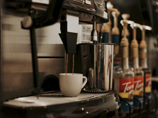 A coffee machine fills a cup, with various syrup bottles lined up in the background.