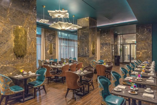 The image shows an elegant restaurant interior with marble walls, teal chairs, and tables set for dining, under stylish light fixtures.