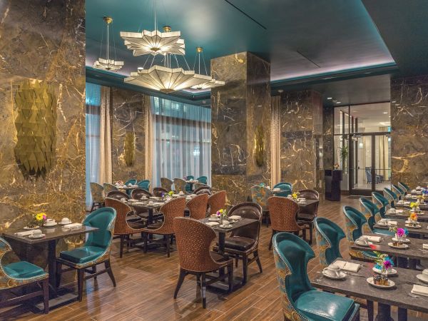 The image shows an elegant restaurant interior with marble walls, teal chairs, and tables set for dining, under stylish light fixtures.