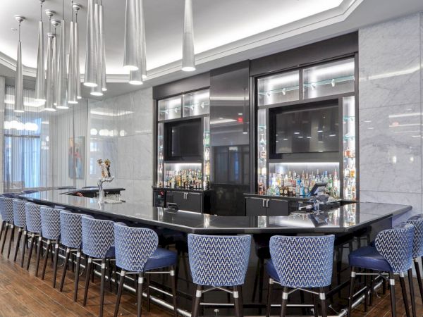 A modern bar with a large countertop, high-backed blue chairs, two TVs, and an array of hanging lights.