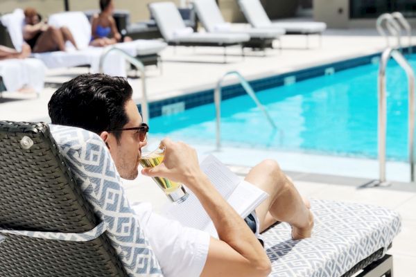 A person relaxes on a lounge chair by a pool, sipping a drink, with more people and lounge chairs in the background.