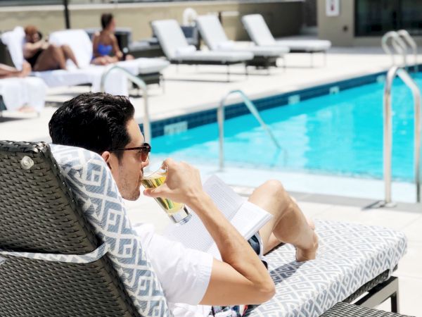 A person relaxes on a lounge chair by a pool, sipping a drink, with more people and lounge chairs in the background.