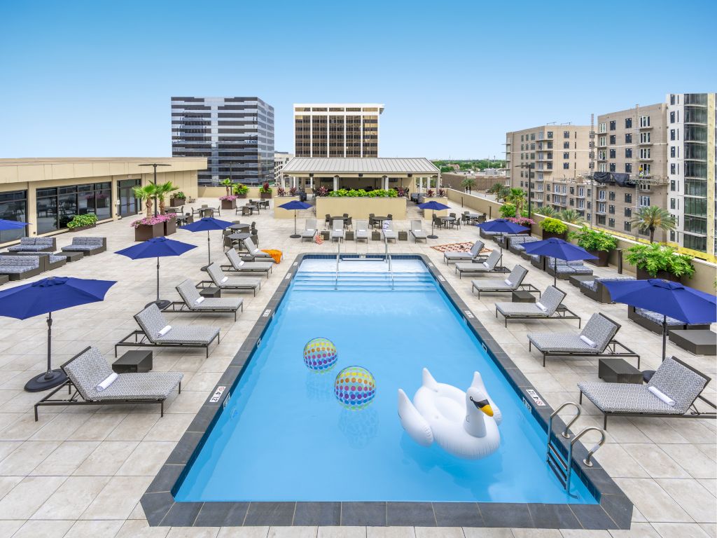 A rooftop pool with a rectangular swimming pool, lounge chairs, blue umbrellas, and inflatable toys.