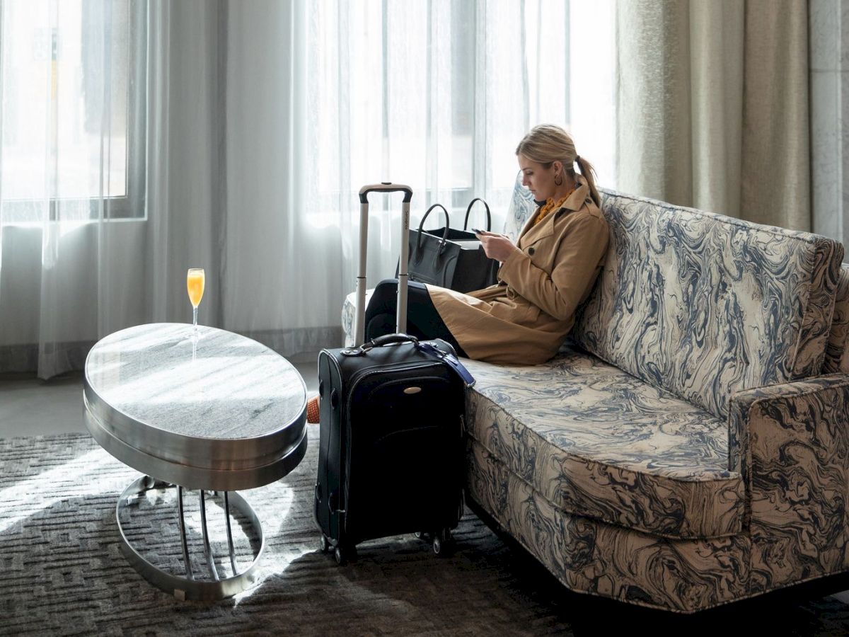 A person sits on a couch, looking at a phone. There's a suitcase nearby and a drink on the table in a well-lit room.