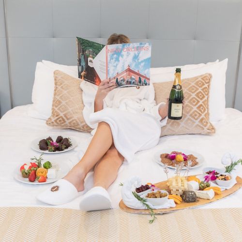 A person in a robe reads a magazine on a bed surrounded by fruit, chocolates, a cheese platter, and champagne. Pillow behind them.