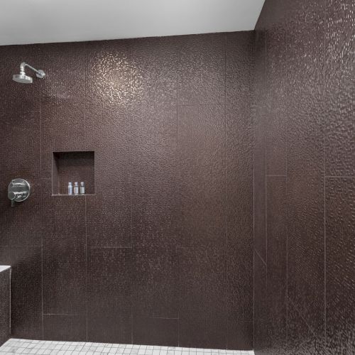 The image shows a modern, brown-tiled shower with a bench, showerhead, handheld spray, built-in shelf, and sleek chrome fixtures.