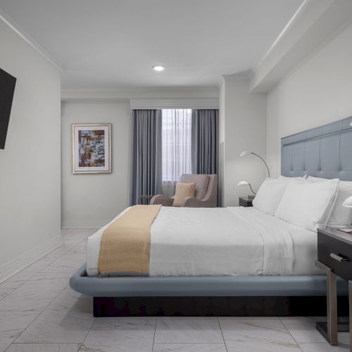 A modern bedroom with a bed, nightstands, a chair, and a wall-mounted TV. Gray and white tones dominate the clean, minimal design.