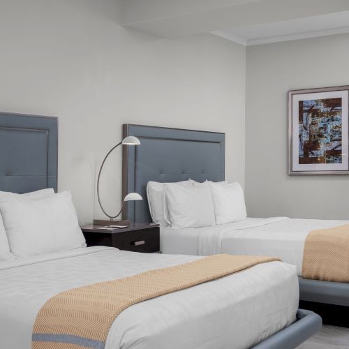 The image shows a hotel room with two beds, a nightstand, a lamp, a wall painting, and a wall light fixture.