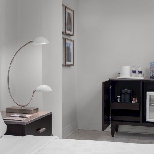 A hotel room corner with a bed, lamp, nightstand, minibar with fridge, coffee maker, bottles, and art on the walls.