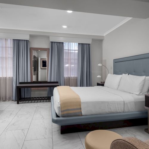 The image shows a modern bedroom with a bed, armchair, large TV, two windows with curtains, and a small table.