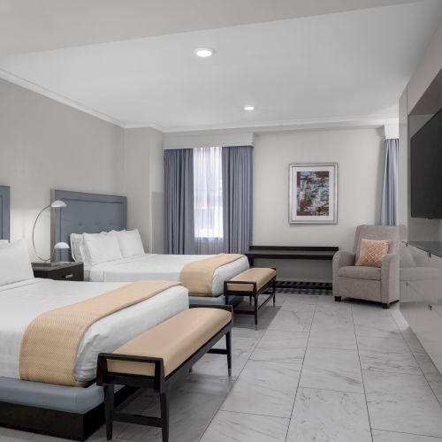 The image shows a modern hotel room with two beds, a TV, seating area, and clean decor, creating a comfortable and inviting atmosphere.