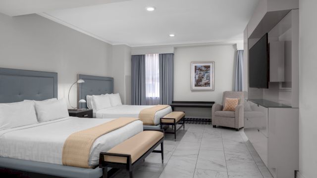 A wide view of a hotel room with two beds, a TV, a chair, and a bench.