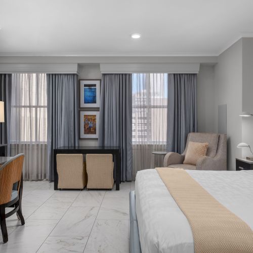 The image shows a modern hotel room with a bed, desk, TV, chair, and large windows with curtains.