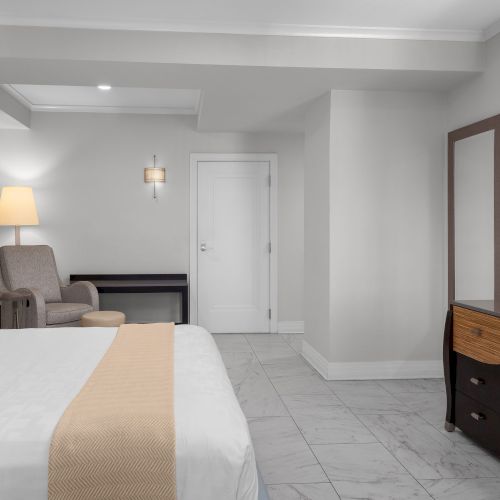 A modern hotel room features a bed, armchair, lamp, flat-screen TV, and a desk, decorated in neutral tones with a minimalist style.