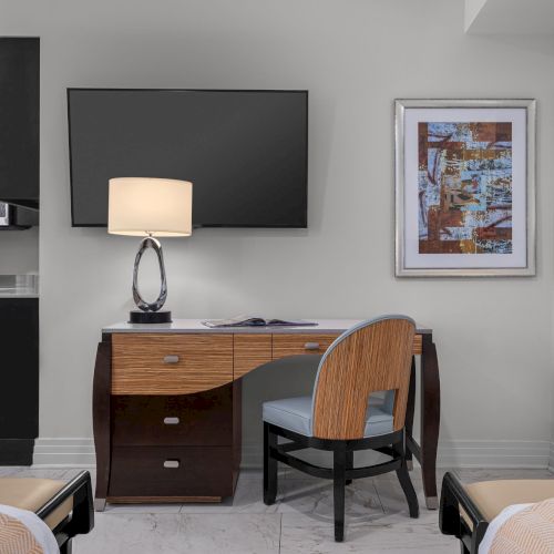 A modern room with a TV, desk, lamp, chair, artwork, and part of two beds visible. A kitchenette is in the corner.