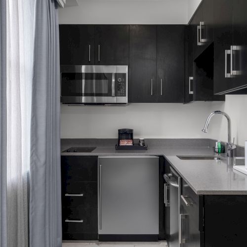 A modern kitchen with dark cabinets, a microwave, sink, coffee maker, and a window with gray curtains.