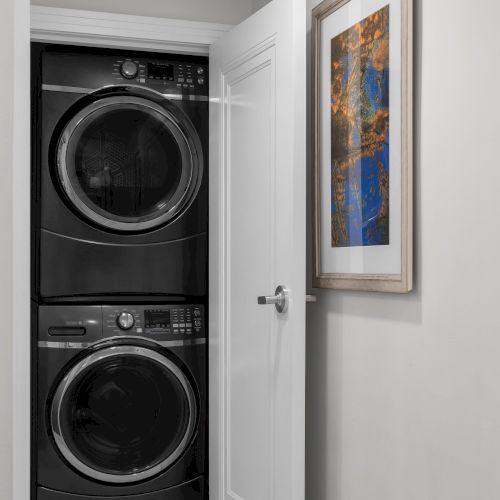 This image shows a stacked washer and dryer inside a closet with an open door and a framed picture on the wall next to it.