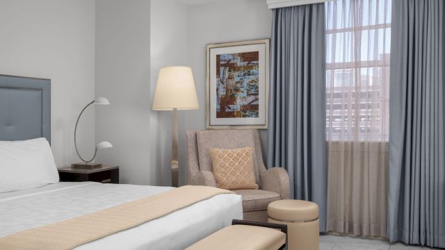 A wide view of a hotel room with a bed, chair, lamp, and window.