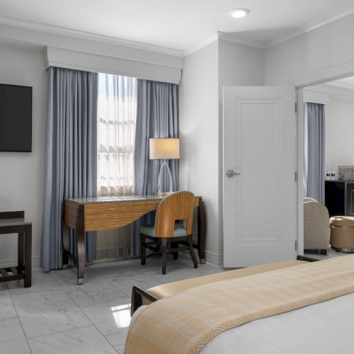 A hotel room with a bed, TV, desk with chair, and lamp, featuring neutral colors and modern decor.