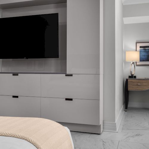 The image shows a modern room with a TV, dresser, bed corner, desk, chair, lamp, and framed pictures on the wall.