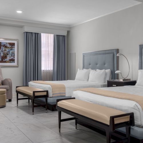 A hotel room with two large beds, stylish headboards, armchair, side table, lamps, and an abstract painting on the wall.