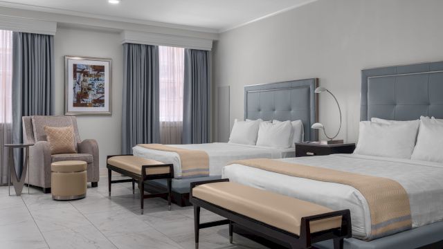 A hotel room with two beds, two benches, a chair, and a table with a lamp.