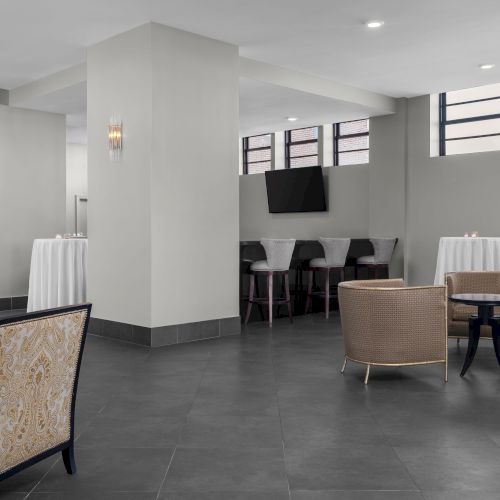 The image shows a modern lounge with chairs, tables, and a wall-mounted TV, featuring a sleek gray and beige color scheme.