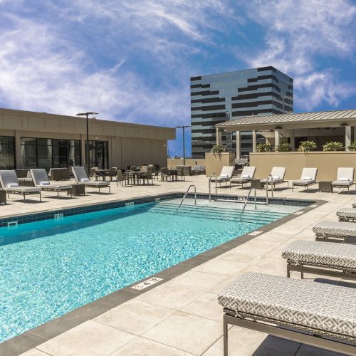 A rooftop pool with lounge chairs, surrounded by buildings, against a blue sky with clouds. The area is clean and inviting for relaxation.