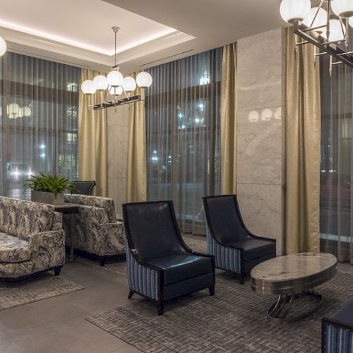The image shows a modern lobby with patterned sofas, dark chairs, marble tables, and elegant lighting, surrounded by large windows and curtains.
