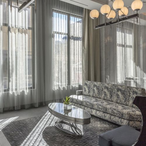 A modern living room with tall windows, sheer curtains, a floral-patterned sofa, oval coffee table, and spherical pendant lights.