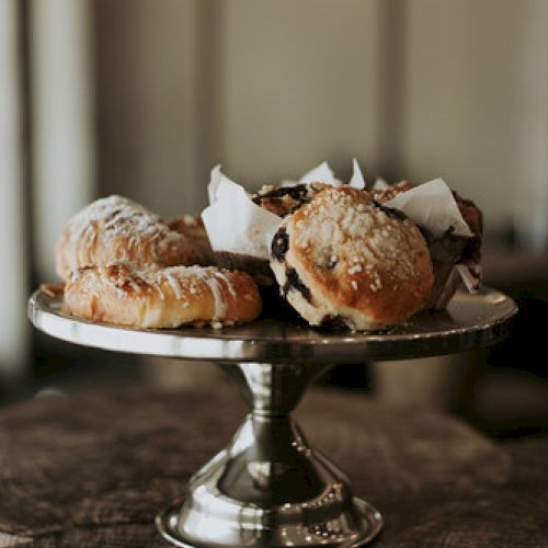 A metal cake stand displays various pastries, including muffins and croissants, atop a rustic wooden surface.
