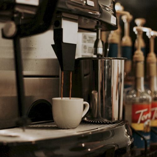 A coffee machine pours coffee into a white mug, surrounded by syrup bottles, on a counter.