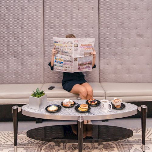 A person reads a newspaper on a beige bench with various pastries and drinks on a modern coffee table in front of them.