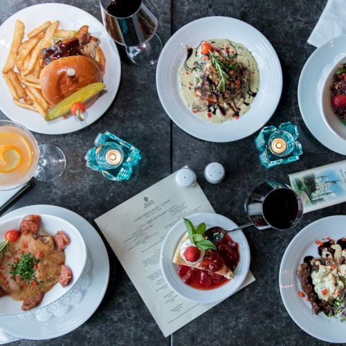 A table with various dishes including a burger, pasta, steak, and desserts, surrounded by drinks, candles, and a menu on a dark surface.