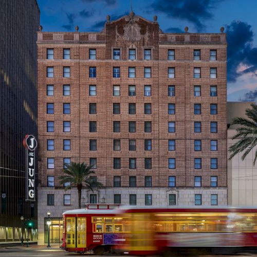A streetcar passes in front of a historic hotel with palm trees and evening sky, featuring a sign reading "Jung" on the building.