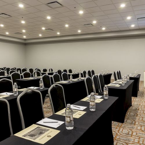 A conference room with rows of black chairs, desks with water bottles, papers, and a distant podium under bright ceiling lights.