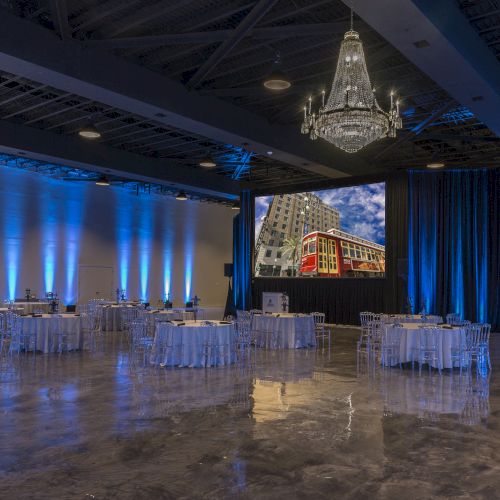 An elegant hall with chandeliers, blue lighting, round tables, and a large screen displaying a street scene with a red trolley.