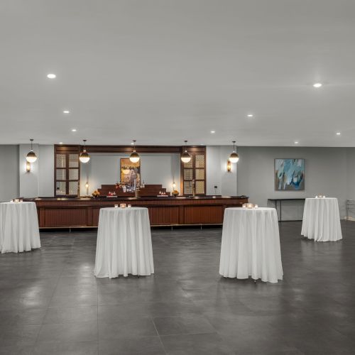 The image shows a modern, elegant event space with a bar, cocktail tables draped in white cloth, and ambient lighting.