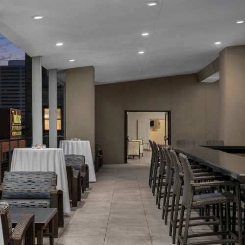 An outdoor patio with a bar, chairs, and tables is shown, with city lights visible in the background at dusk.