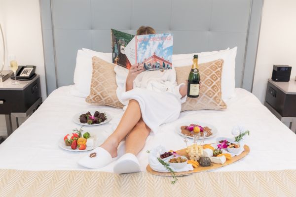 A person in a robe relaxes on a bed with a magazine, holding a champagne bottle, surrounded by snacks and fruit platters.